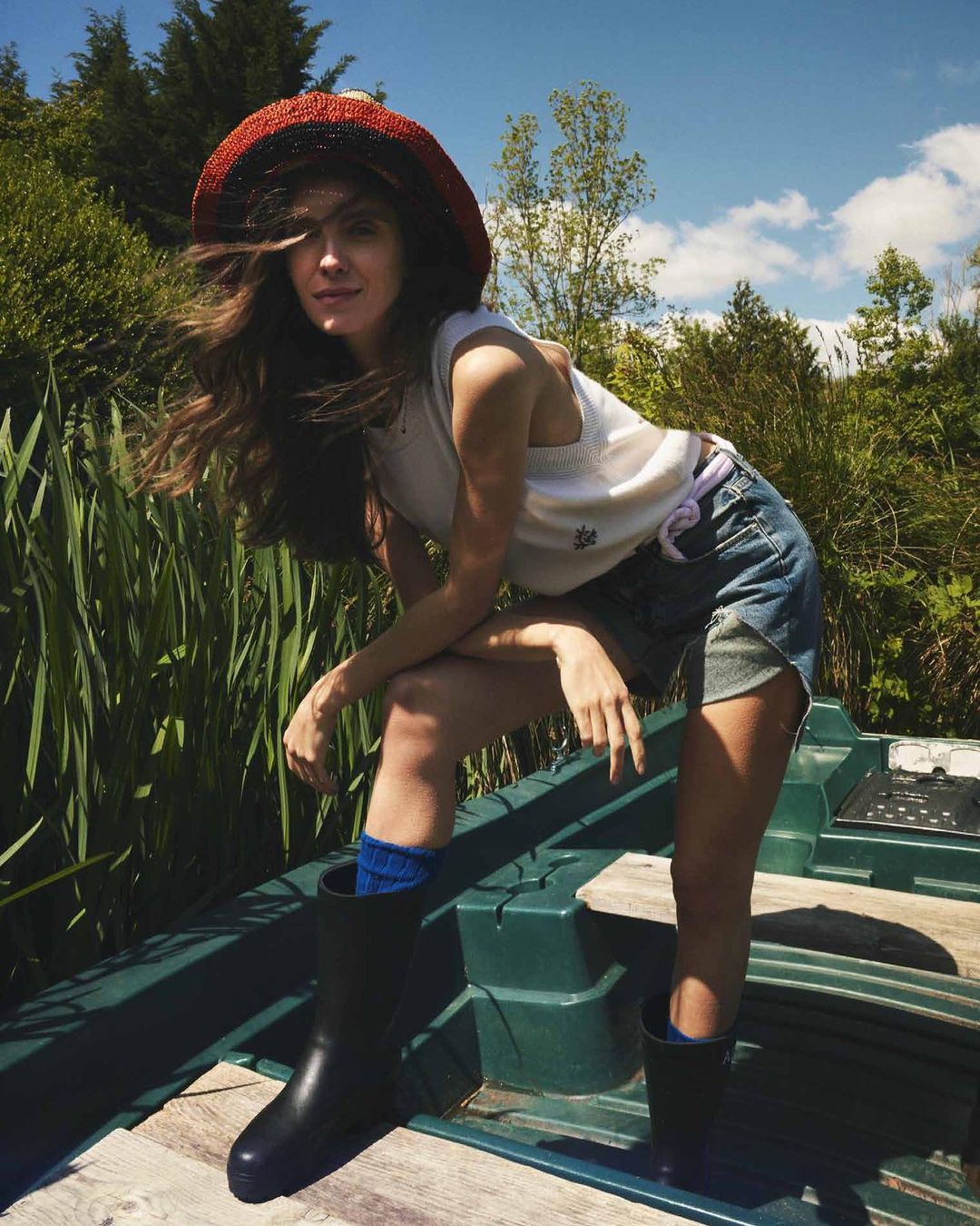A woman in a hat, tank top, and denim shorts sits on a boat in a lush, green outdoor setting under a partly cloudy sky.
