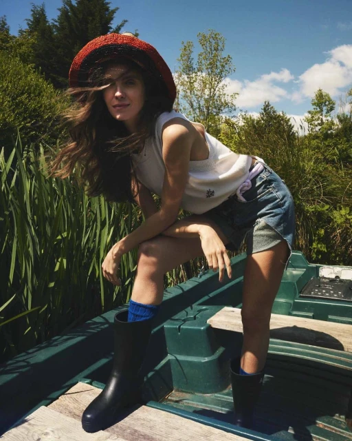A woman in a hat, tank top, and denim shorts sits on a boat in a lush, green outdoor setting under a partly cloudy sky.