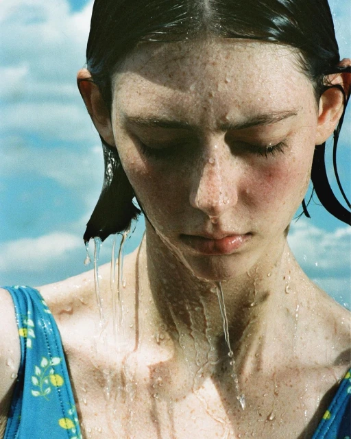 Close-up of a young woman with freckles and wet hair, water dripping from her face, wearing a blue floral swimsuit against a blue sky.