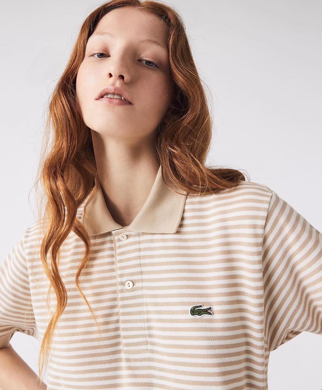 A fair-skinned young woman with red hair models a striped tan and white Lacoste polo shirt in a bright, neutral studio setting.