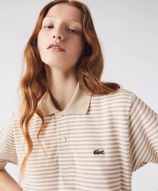 A fair-skinned young woman with red hair models a striped tan and white Lacoste polo shirt in a bright, neutral studio setting.