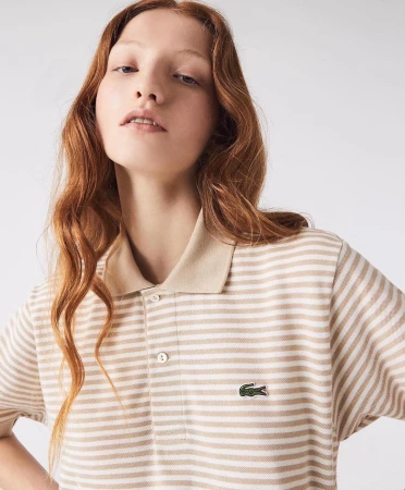 A fair-skinned young woman with red hair models a striped tan and white Lacoste polo shirt in a bright, neutral studio setting.