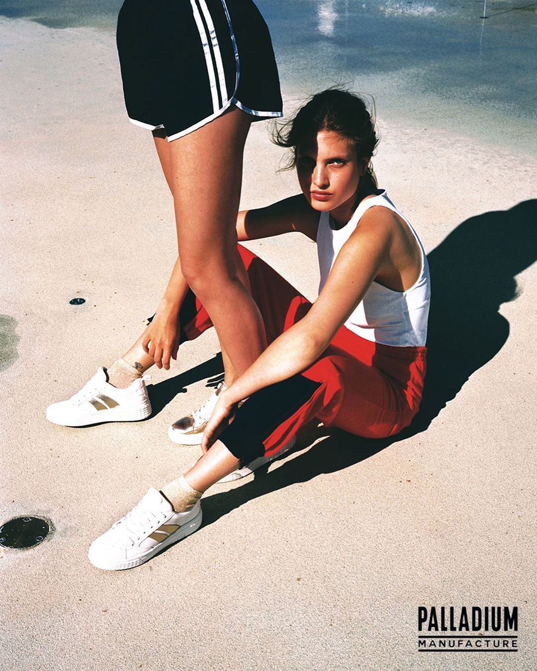Two women in activewear sit on a light-beige concrete surface with a water feature in the background, both wearing white sneakers.