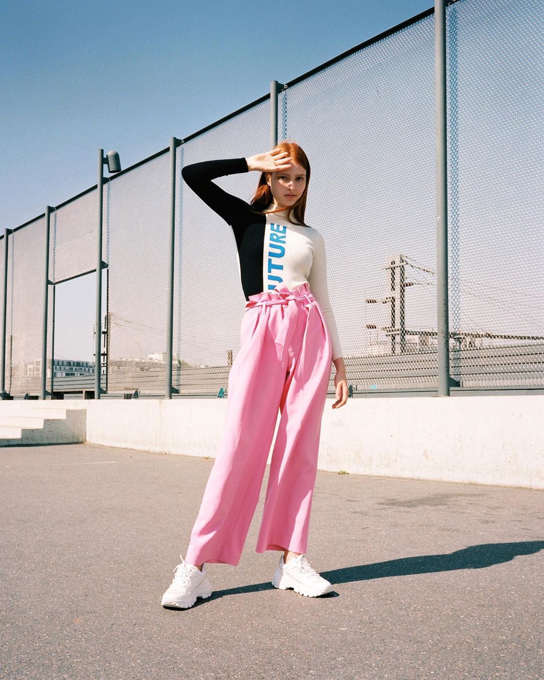 A woman in pink pants and a black and white top poses against a blue sky and tennis court. She is shielding her eyes.