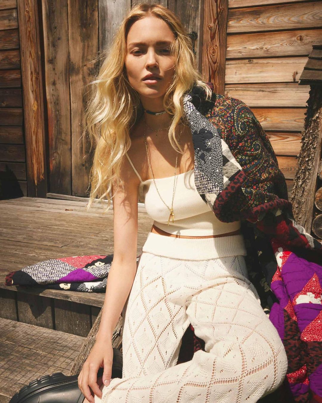 Blonde woman in knit pants and top poses on wooden stairs, draped with a patterned coat, bathed in soft sunlight near a wood cabin.