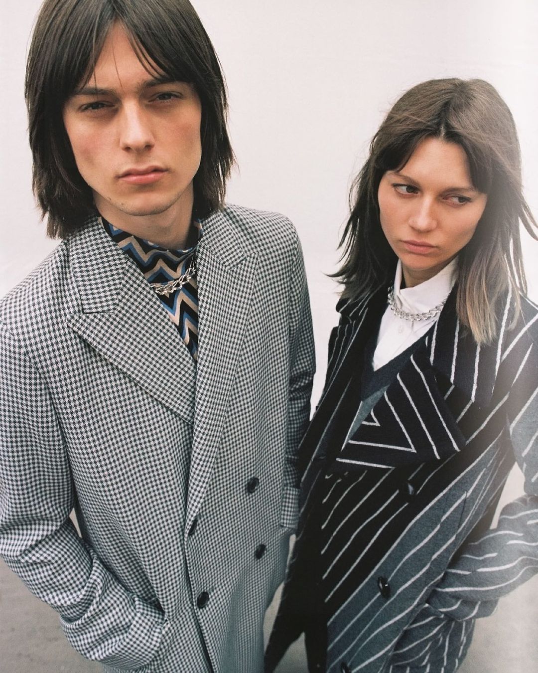 Two models, one male and one female, pose in stylish patterned overcoats and silver chain necklaces against a plain neutral backdrop.