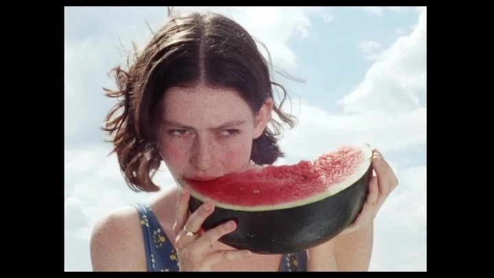 Close-up of a young woman with short brown hair eating watermelon against a light blue sky with white clouds, enjoying a fresh snack.