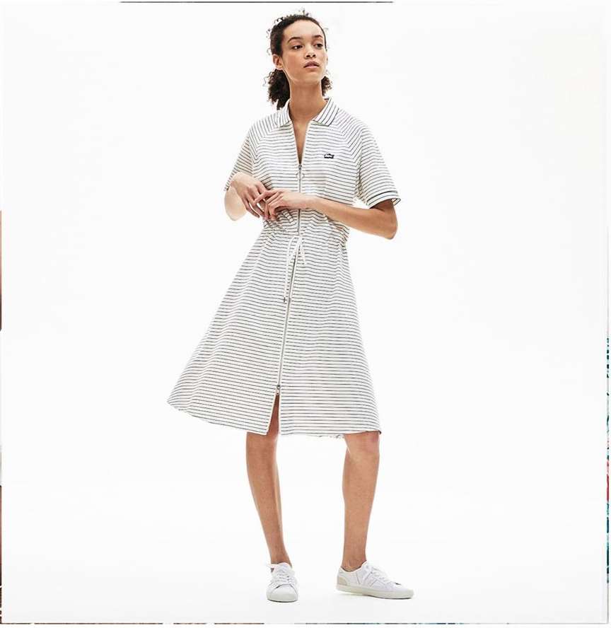 A woman wears a Lacoste striped, collared shirt dress with a zipper and a tie waist, paired with white sneakers against a white backdrop.