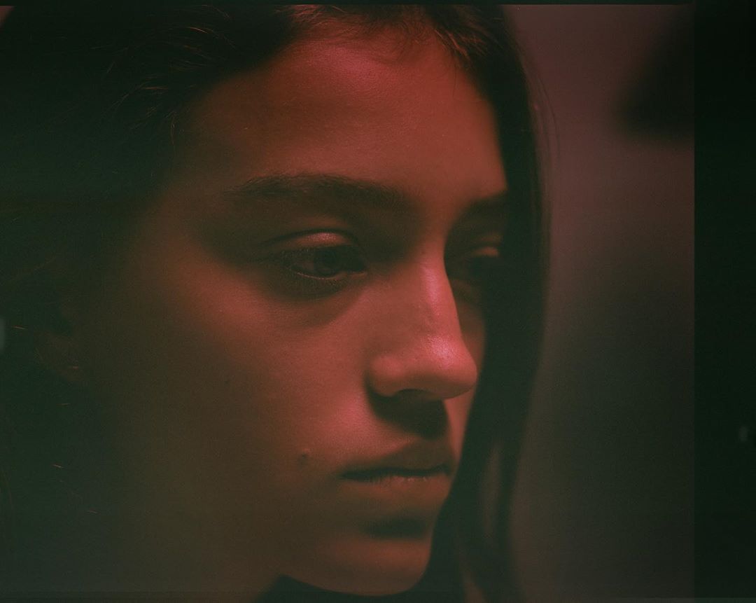Portrait of a young woman with dark hair in a close-up shot, displaying a serene facial expression under red-toned lighting in an indoor setting.
