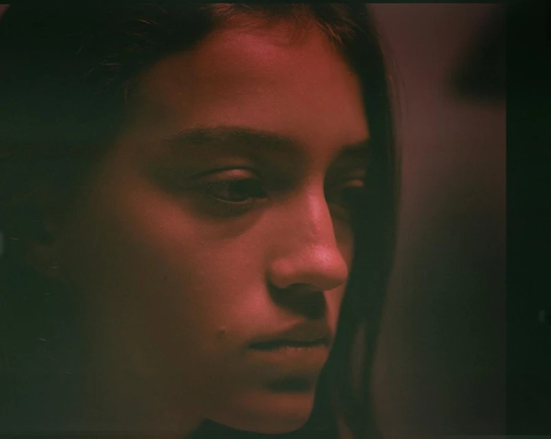Portrait of a young woman with dark hair in a close-up shot, displaying a serene facial expression under red-toned lighting in an indoor setting.