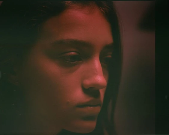 Portrait of a young woman with dark hair in a close-up shot, displaying a serene facial expression under red-toned lighting in an indoor setting.