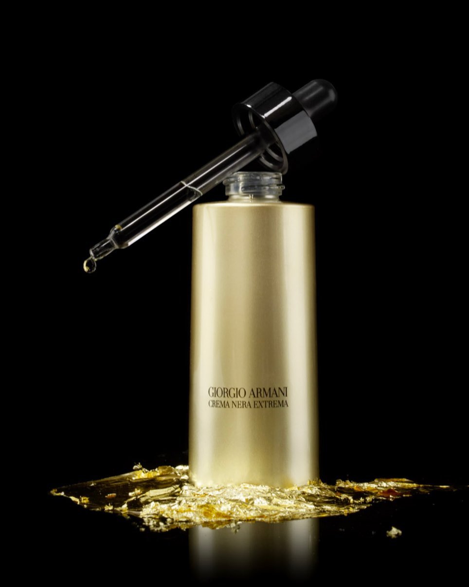 Product shot of Giorgio Armani Crema Nera Extrema skincare serum bottle with dropper, liquid, and gold flakes on a black background.