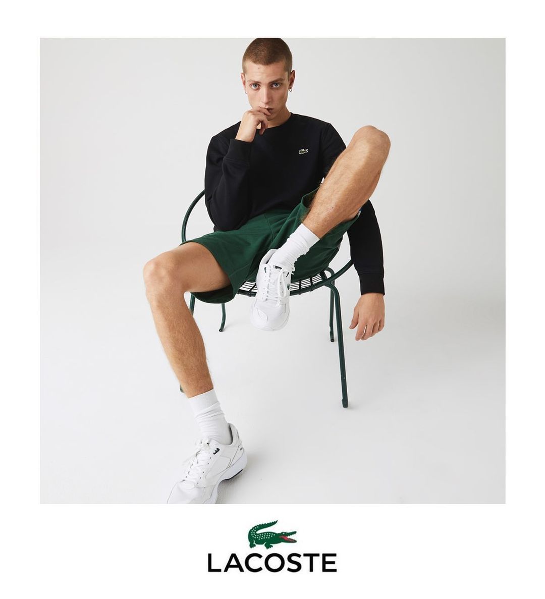 Young man in a black sweatshirt and green shorts seated on a wire chair against a white background by Max Vigato