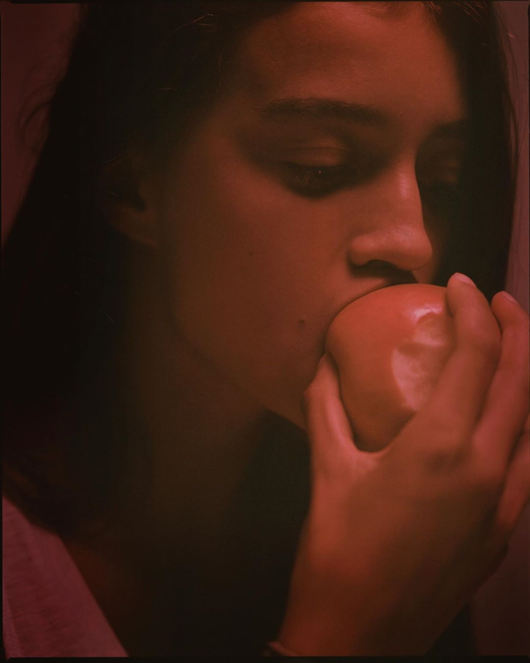 Close-up of a woman biting into a red apple, her hand holding the fruit, bathed in red lighting in an indoor setting.