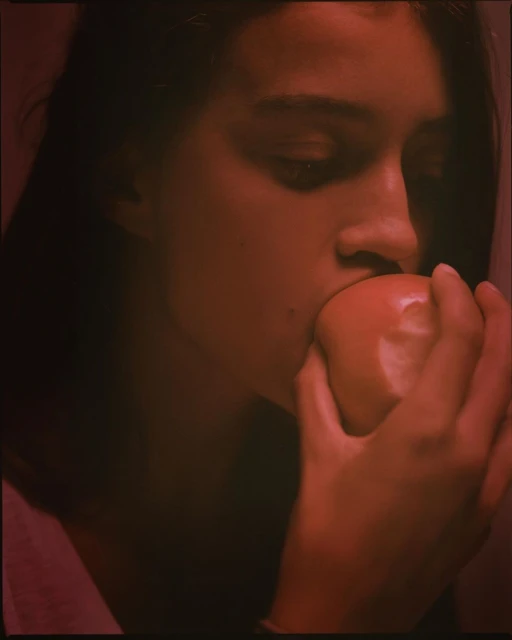 Close-up of a woman biting into a red apple, her hand holding the fruit, bathed in red lighting in an indoor setting.