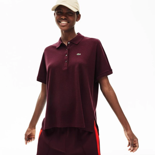A woman with a cap is smiling while posing in a maroon Lacoste polo shirt and matching skirt against a bright white backdrop.