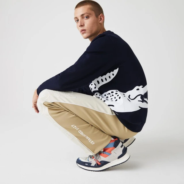 A male model crouches in a studio wearing a navy blue Lacoste sweatshirt with a large white crocodile logo, cream and beige pants, and colorful sneakers.