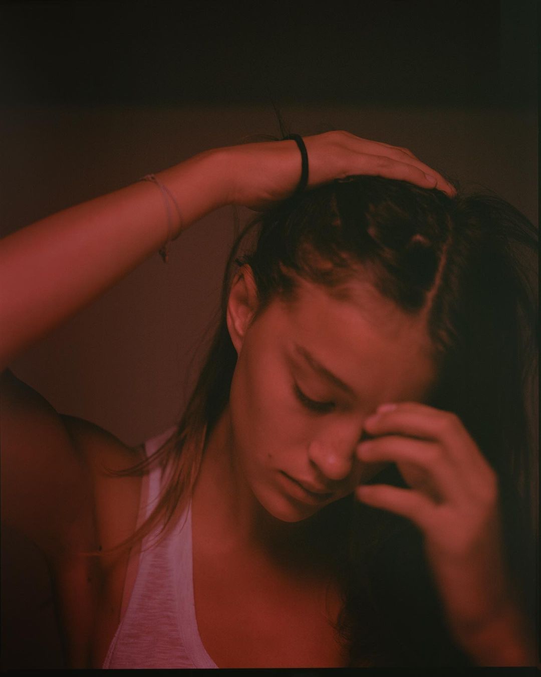 Close-up portrait of a young woman with dark hair, wearing a white tank top, with her hands on her head, lit with a red hue.