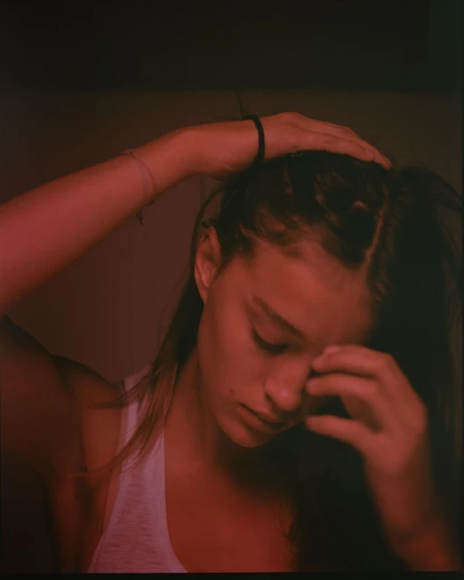 Close-up portrait of a young woman with dark hair, wearing a white tank top, with her hands on her head, lit with a red hue.