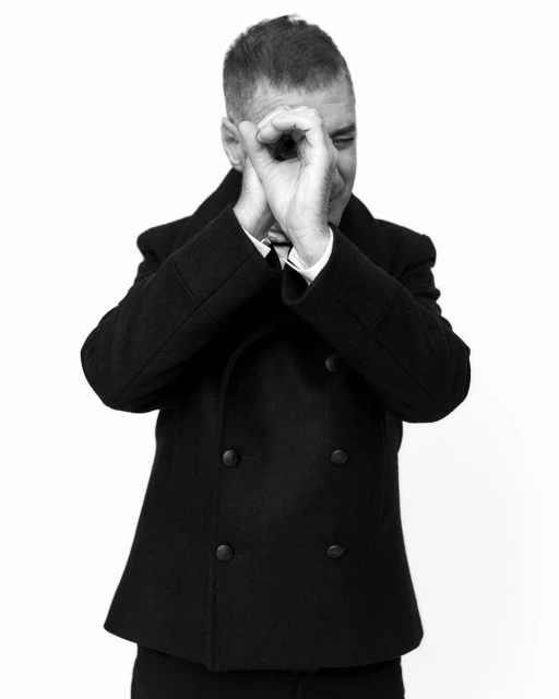 Monochrome portrait of an adult male wearing a peacoat, making a circle shape with his hands, set against a plain white backdrop.