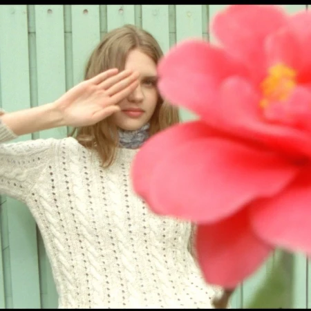 Young woman in a textured white sweater holding her hand to her eye, partially obscured by a large bright pink flower.