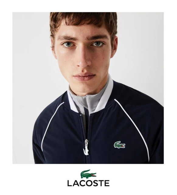 Close-up of a young adult male wearing a navy Lacoste bomber jacket with white trim and a gray collared shirt against a neutral backdrop.