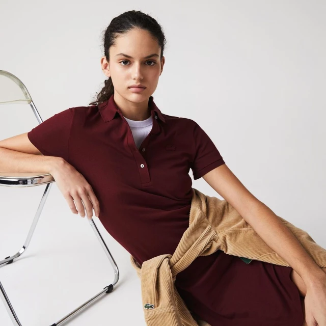 Adult woman in burgundy polo dress leans on clear chair with beige sweater in a bright studio with a neutral background.