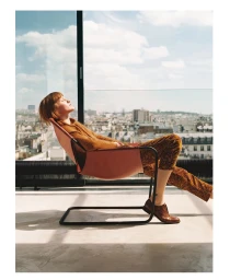 Woman with short red hair reclining in a pink lounge chair by a large window overlooking a city skyline, photographed by Max Vigato.