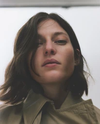 Low-angle portrait of a woman with dark shoulder-length hair wearing a khaki jacket against a plain white background