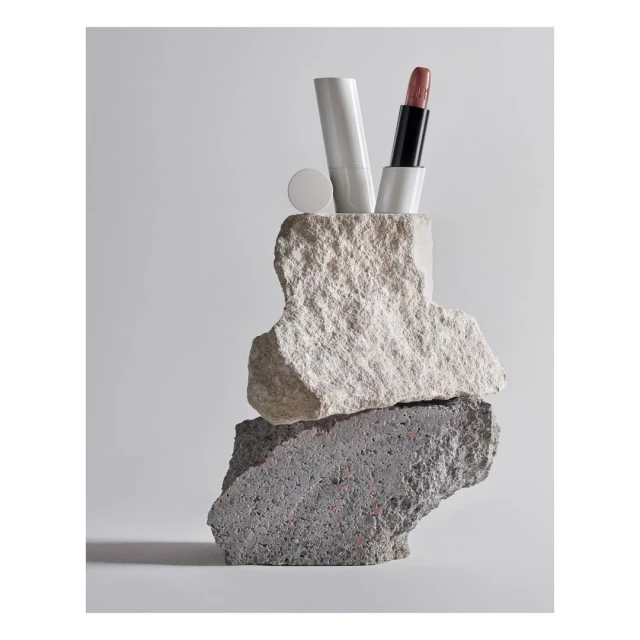 Cosmetic product photography featuring lipstick tube and white container balanced on rough stone, showcasing beauty and textures.