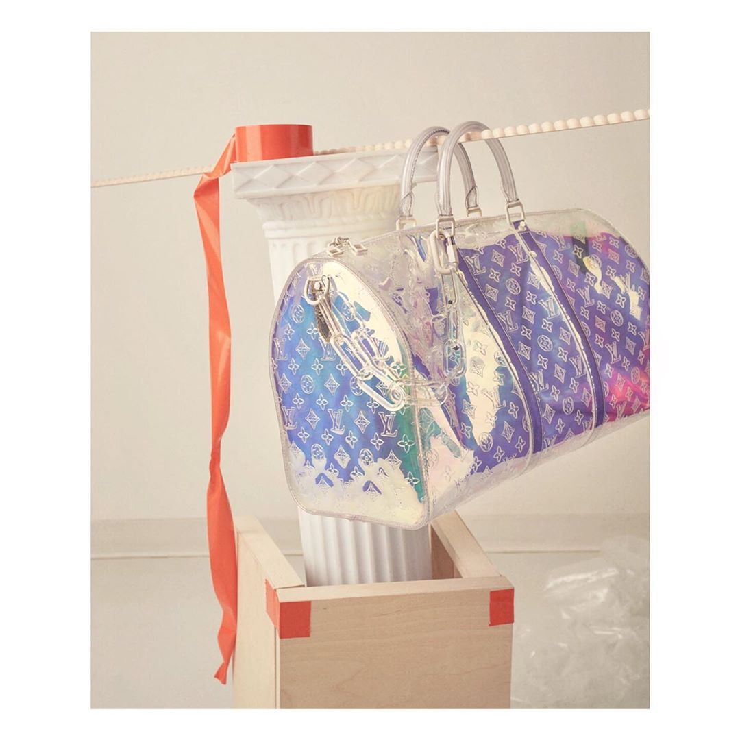 Studio shot of a clear Louis Vuitton handbag hanging above a wooden box, accented by a white column and an orange ribbon.