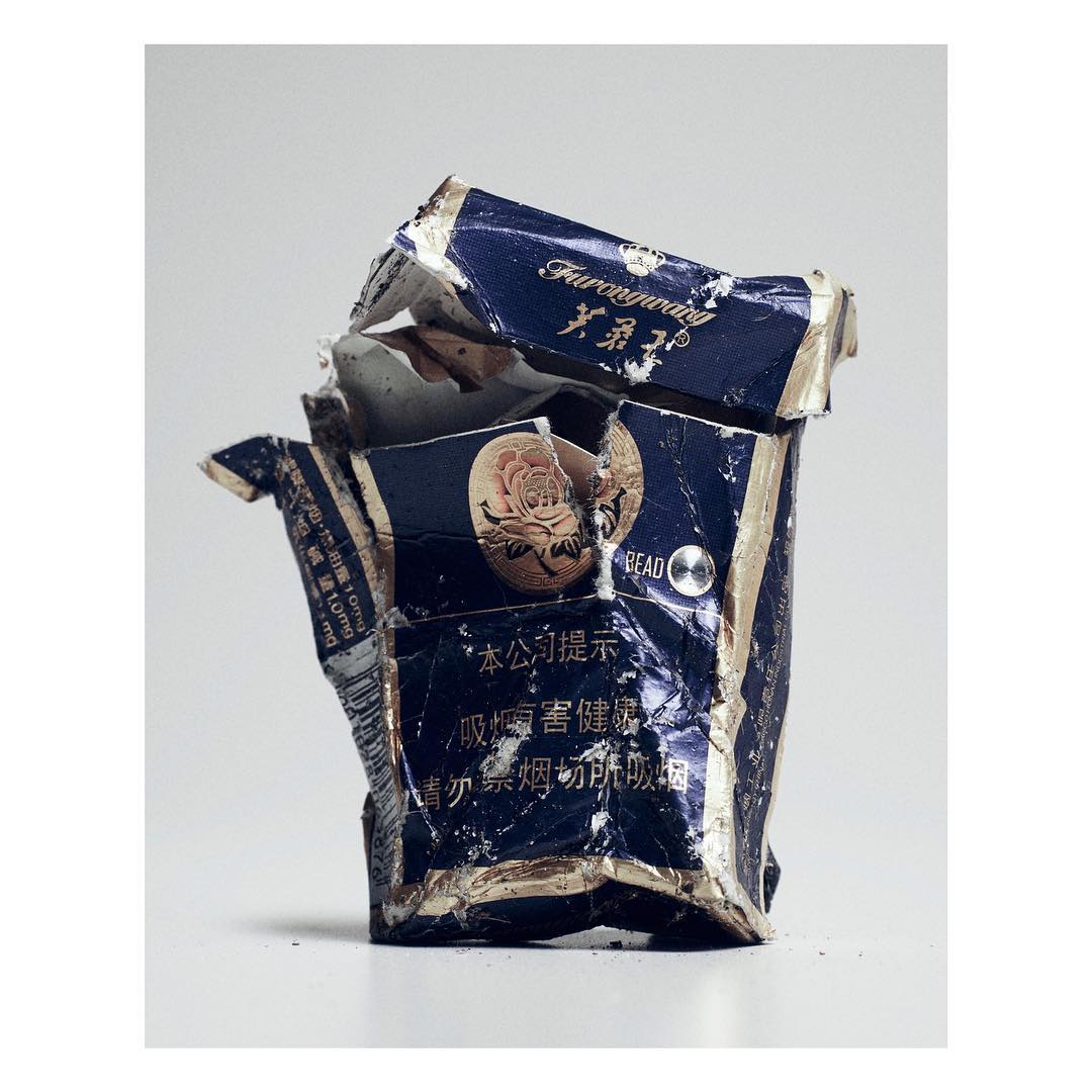 Crushed blue and gold cigarette packet with Chinese writing, resting on a white surface with a soft, high-key lighting setup.