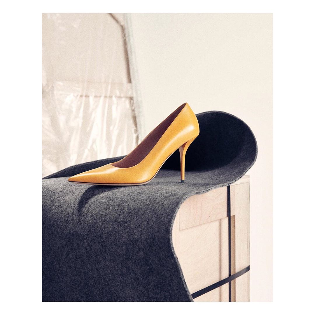 Studio shot of a yellow leather high-heel pump shoe on a gray fabric surface draped over a light wooden box against a white backdrop.
