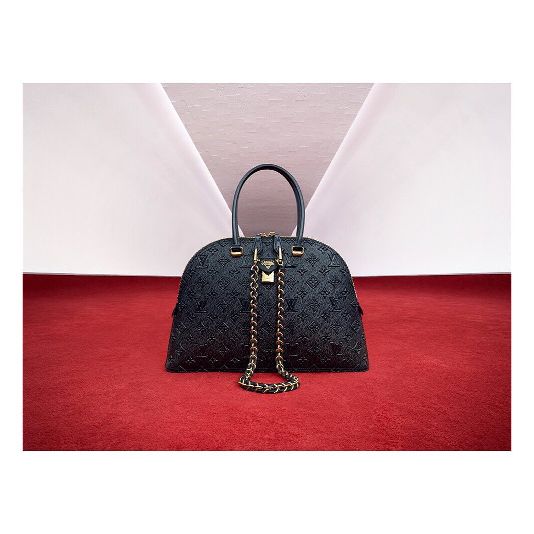 Black leather dome handbag with gold chain and hardware, set against a red carpet and white backdrop in a studio setting.
