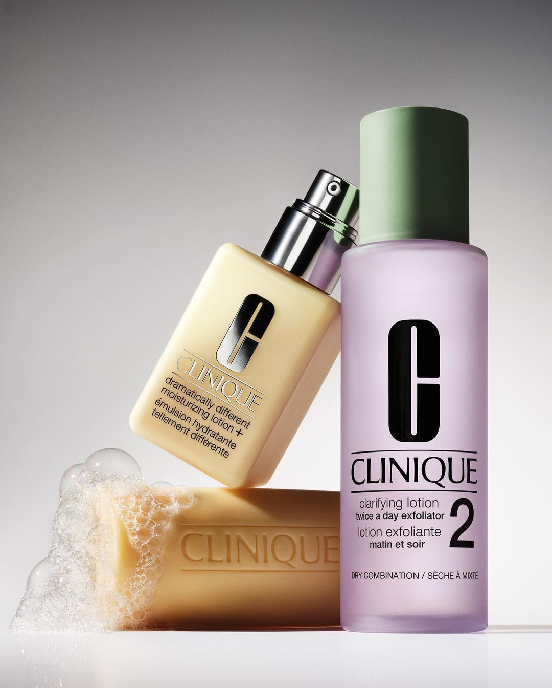 Close-up shot of Clinique skincare products including lotion, exfoliator, and soap, against a gradient gray background. Beauty advertising.