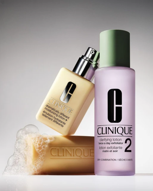 Close-up shot of Clinique skincare products including lotion, exfoliator, and soap, against a gradient gray background. Beauty advertising.