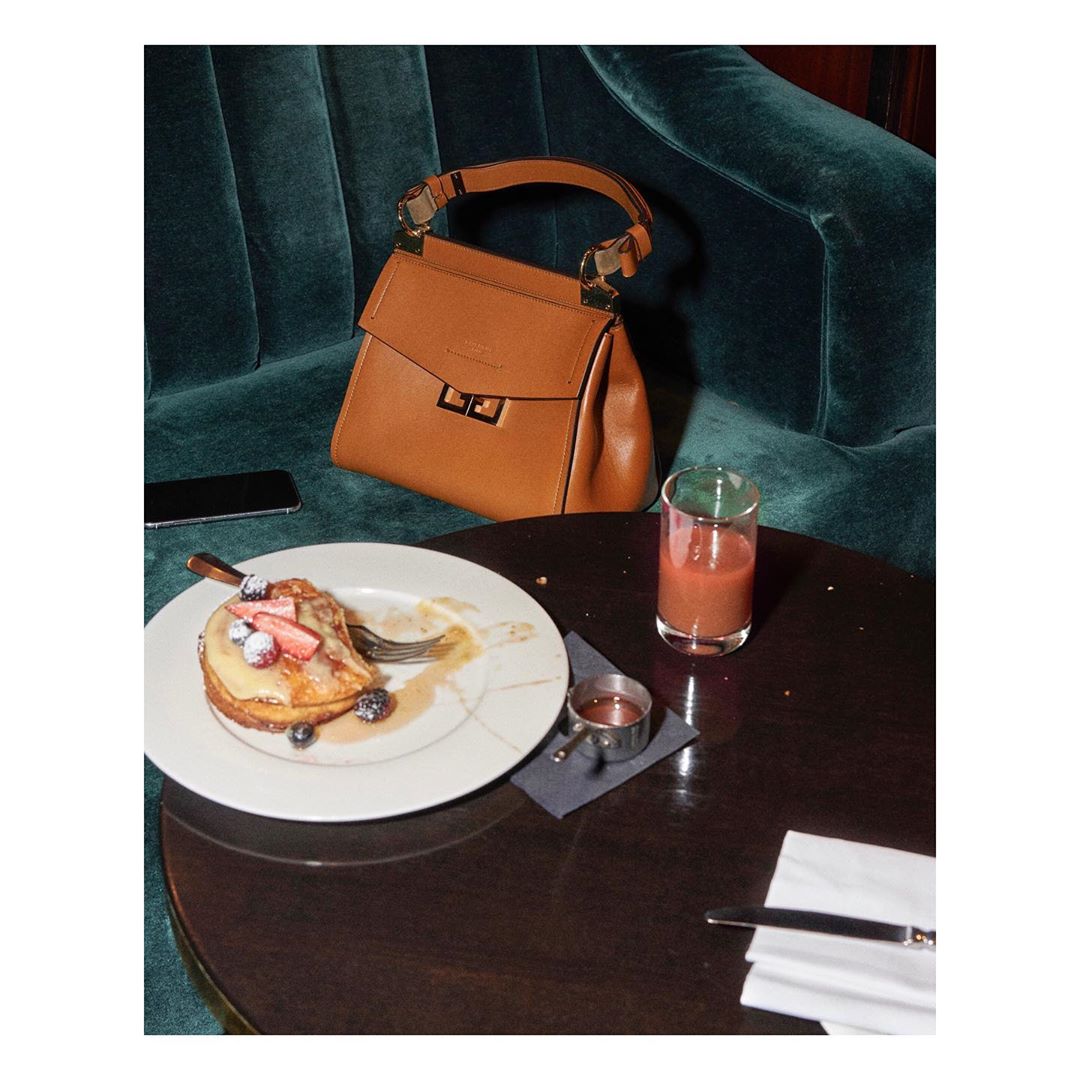 Tan designer purse on a blue velvet couch with a plate of pancakes with fruit and a beverage on a round wooden table.