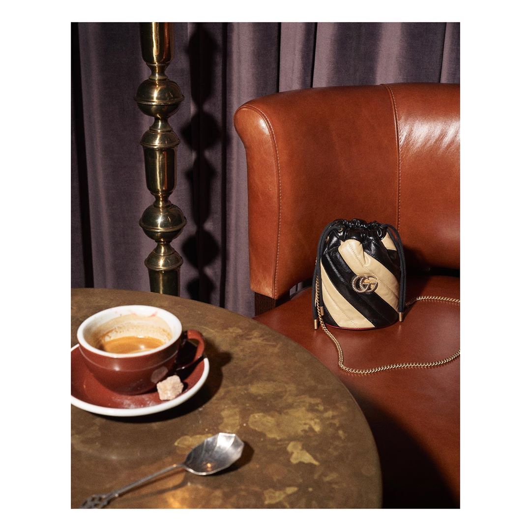 Still life shot of a Gucci purse on a chair next to a table with a cup of coffee, a spoon, and sugar cubes in an indoor setting.