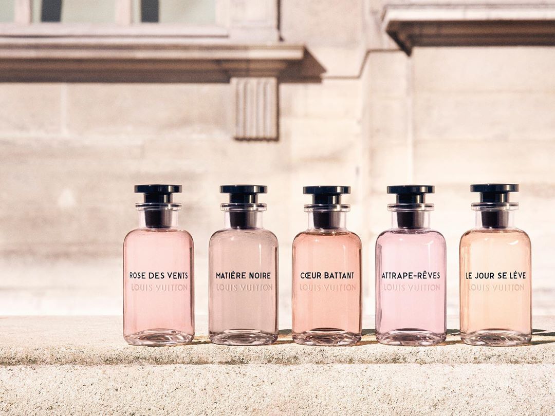Five glass perfume bottles with black caps lined up in a row on a stone ledge outside a classic building.