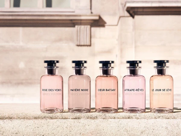 Five glass perfume bottles with black caps lined up in a row on a stone ledge outside a classic building.