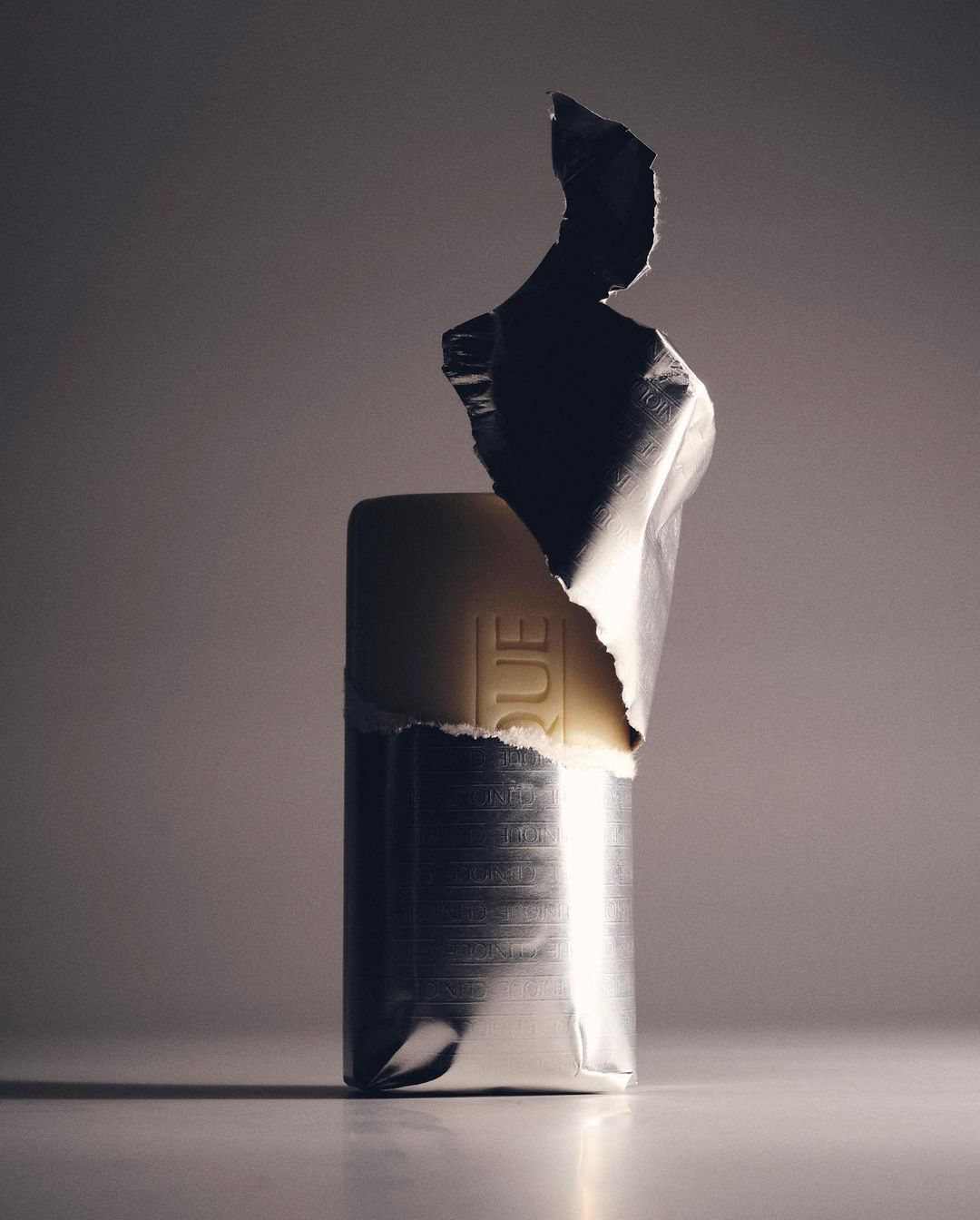 Wrapped bar with silver foil packaging illuminated in studio setting, casting soft shadows on a light surface against a grey background.
