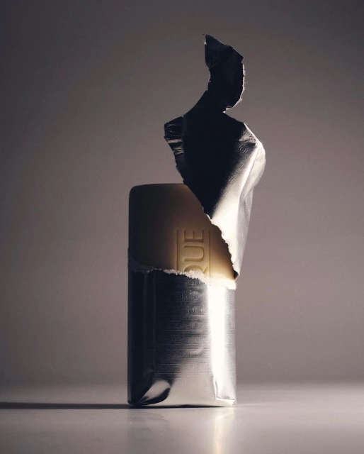 Wrapped bar with silver foil packaging illuminated in studio setting, casting soft shadows on a light surface against a grey background.