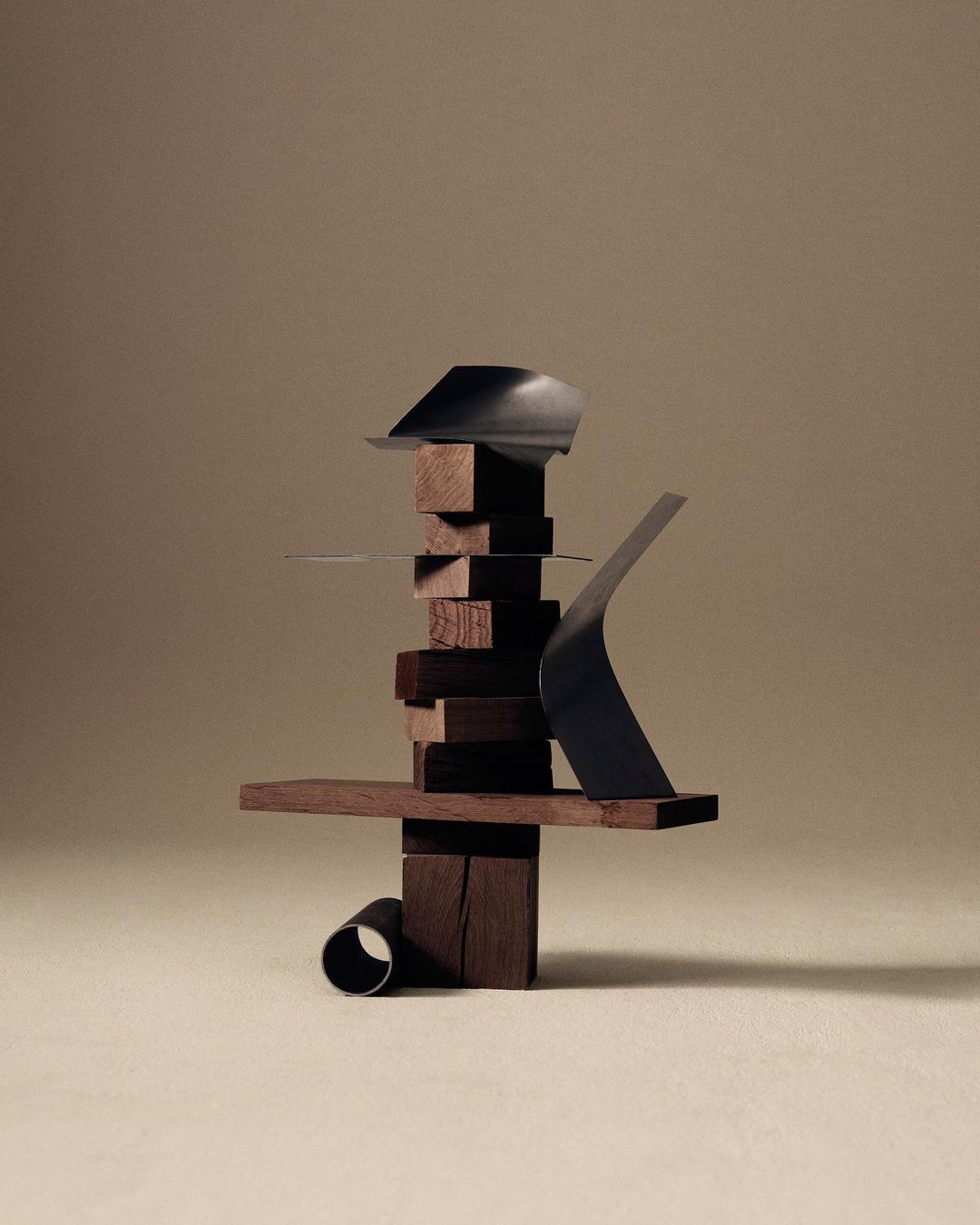 Abstract sculptural composition with dark brown stacked wooden blocks and dark metallic elements on a tan background in studio lighting.
