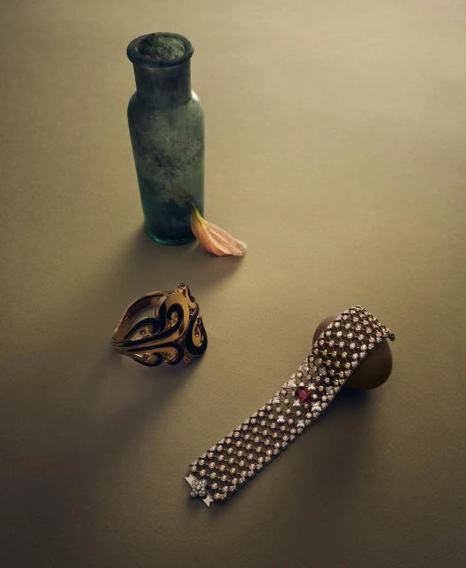 Still life shot of gold jewelry, including a ring with black enamel detailing and a bracelet with white gemstones, next to a glass bottle.