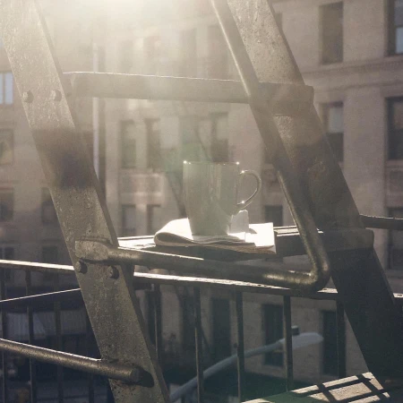 Ceramic mug and newspaper resting on a metal fire escape platform with soft morning sunlight.
