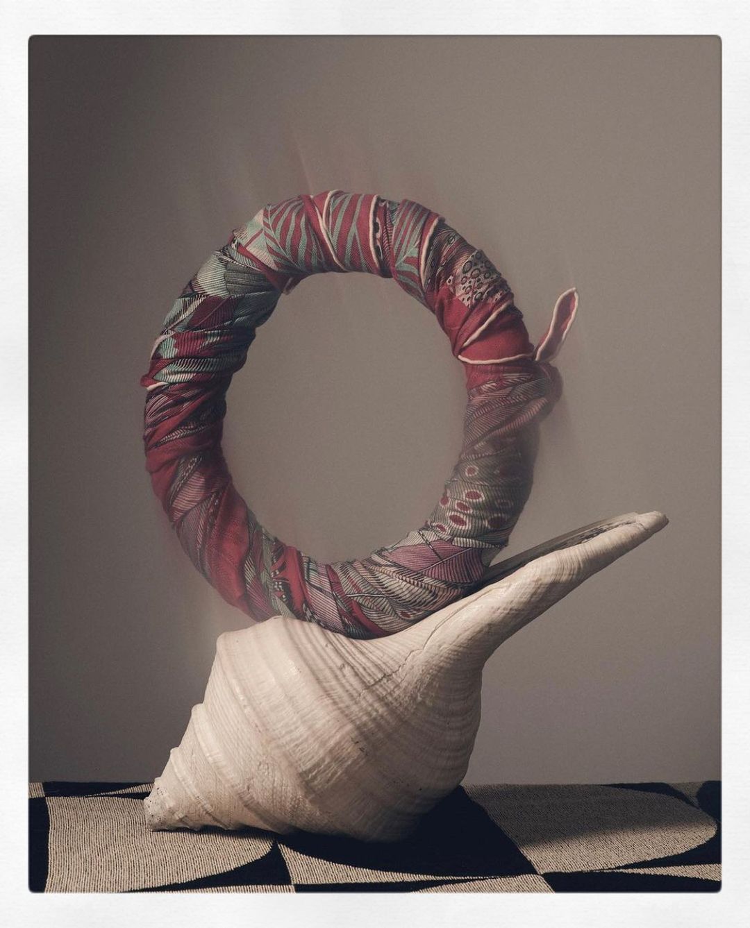Fabric-wrapped ring rests on a large conch shell atop a patterned rug indoors in a still life, showcasing art and decor.