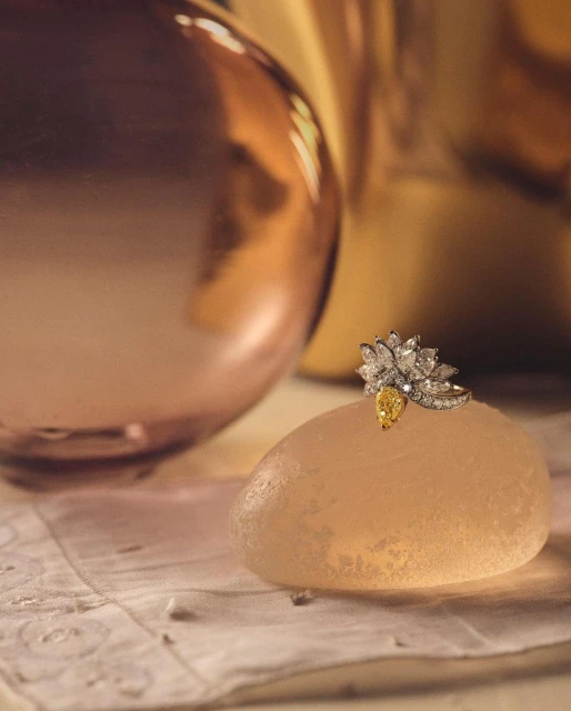 Close-up studio shot of a diamond ring with a yellow gem on a textured stone, with blurred rose gold and gold vases in background.