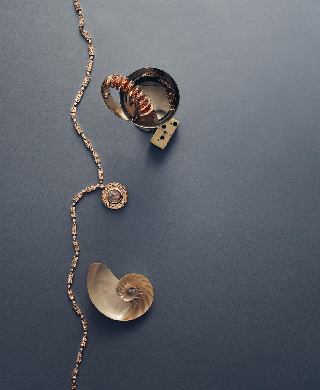 Silver cup, domino, shell, and gold necklace arranged on a dark blue surface in a flat lay composition. Fashion and still life.