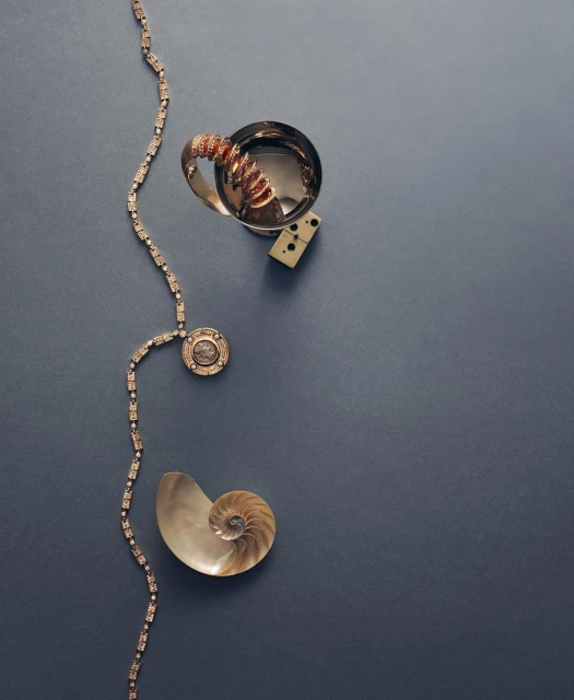 Silver cup, domino, shell, and gold necklace arranged on a dark blue surface in a flat lay composition. Fashion and still life.
