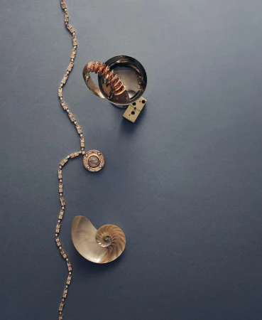 Silver cup, domino, shell, and gold necklace arranged on a dark blue surface in a flat lay composition. Fashion and still life.